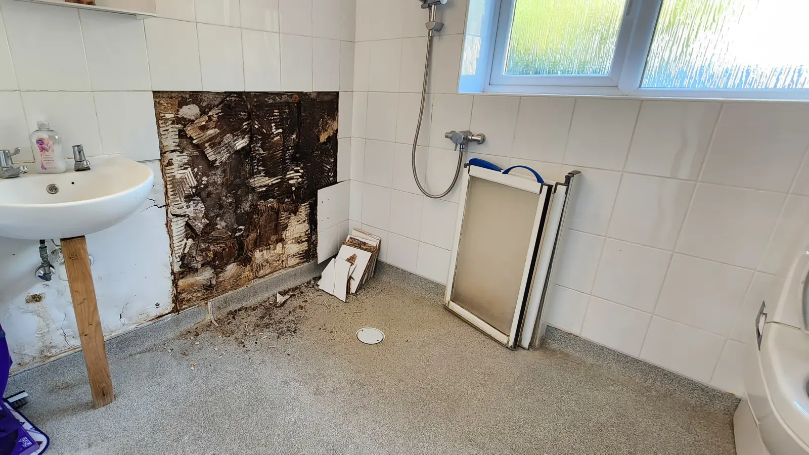 Bathroom wall with a section of tiles removed, exposing the substrate beside the sink and shower